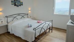 A bedroom with a metal bed frame, white bedding, a bedside table with a lamp, and a window with a view of the landscape.