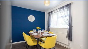 A dining area with a blue accent wall, a round mirror, yellow chairs, and a table set for four. - Fully kitted, 4BR w/Parking, private Garden, Wi-Fi,Near City, Stadiums & Airport (Manchester)