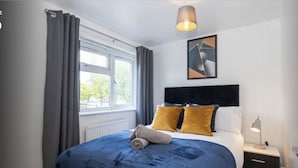 A bedroom with a large bed, a headboard, a window with curtains, a wall-mounted picture, and a bedside lamp. - Fully kitted, 4BR w/Parking, private Garden, Wi-Fi,Near City, Stadiums & Airport (Manchester)