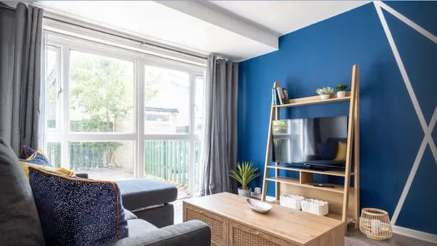 A modern living room with a large window, a blue accent wall, a wooden TV stand, and a grey sofa. - Fully kitted, 4BR w/Parking, private Garden, Wi-Fi,Near City, Stadiums & Airport (Manchester)