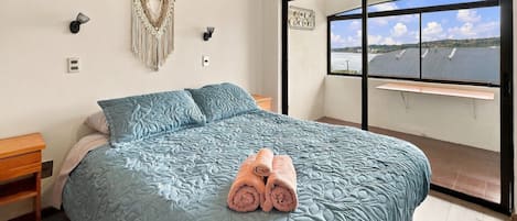 Un dormitorio con una cama grande, una mesita de noche, una lámpara de pared y vistas al exterior a través de un ventanal.