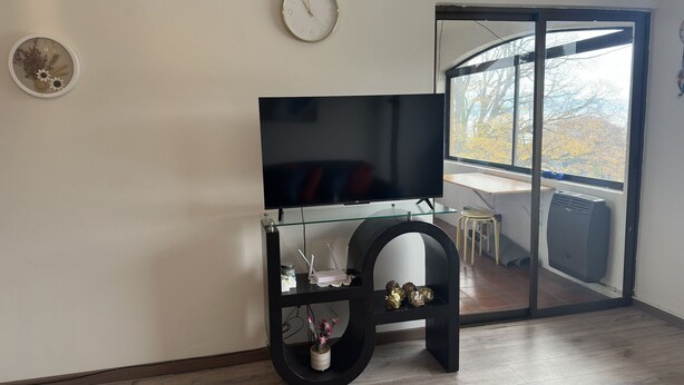A living room with a flat-screen TV on a glass table, a wall clock, and a sliding glass door leading to a balcony. - Oceana Suites Ensenada Villarrica (Villarrica)