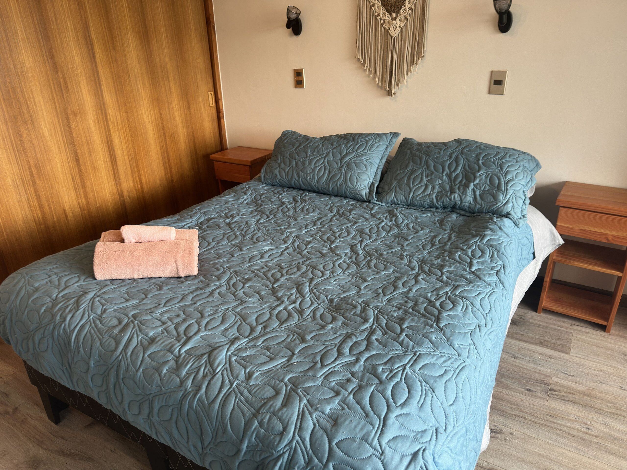Una cama con colcha estampada con motivos de hojas, una mesita de noche de madera, un adorno de macramé fijado a la pared y suelo de madera.