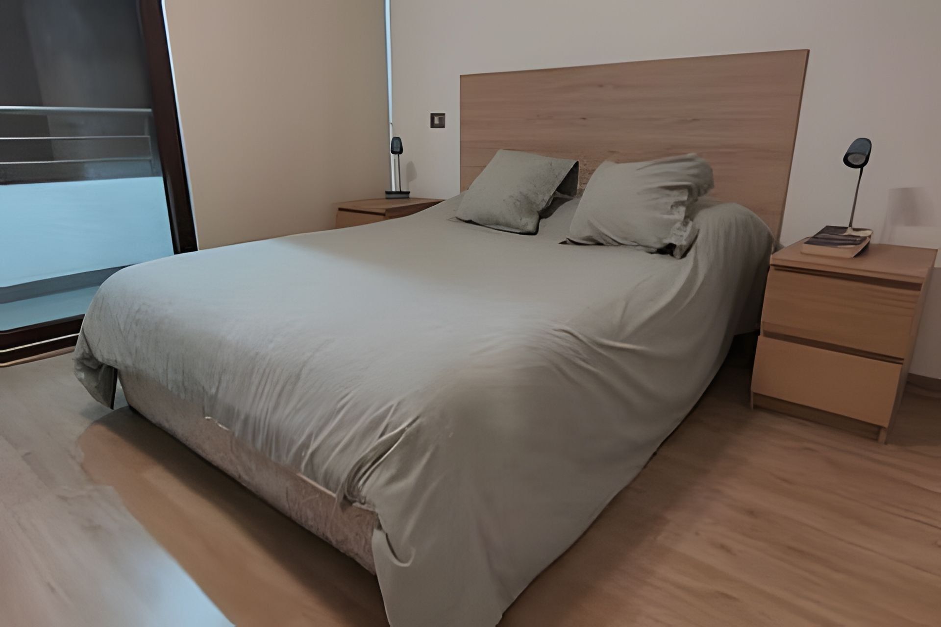 A neatly made bed with a wooden headboard, two bedside tables with lamps, and a large window with blinds.