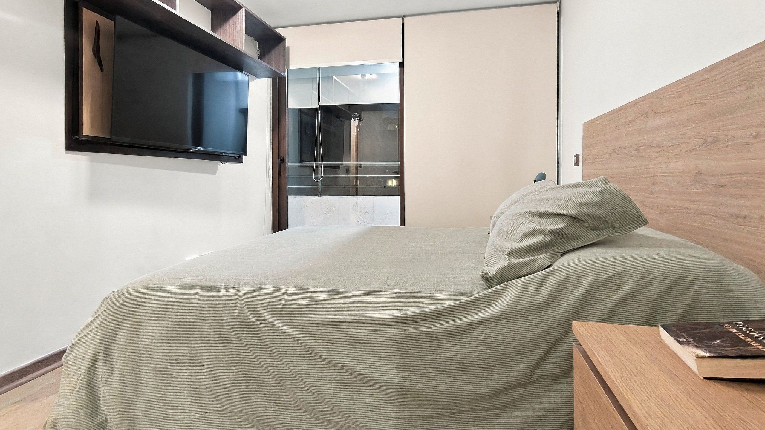 Un dormitorio con cama, mesita de noche con un libro, armario y televisor montado en la pared.