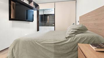 Un dormitorio con cama, mesita de noche con un libro, armario y televisor montado en la pared.