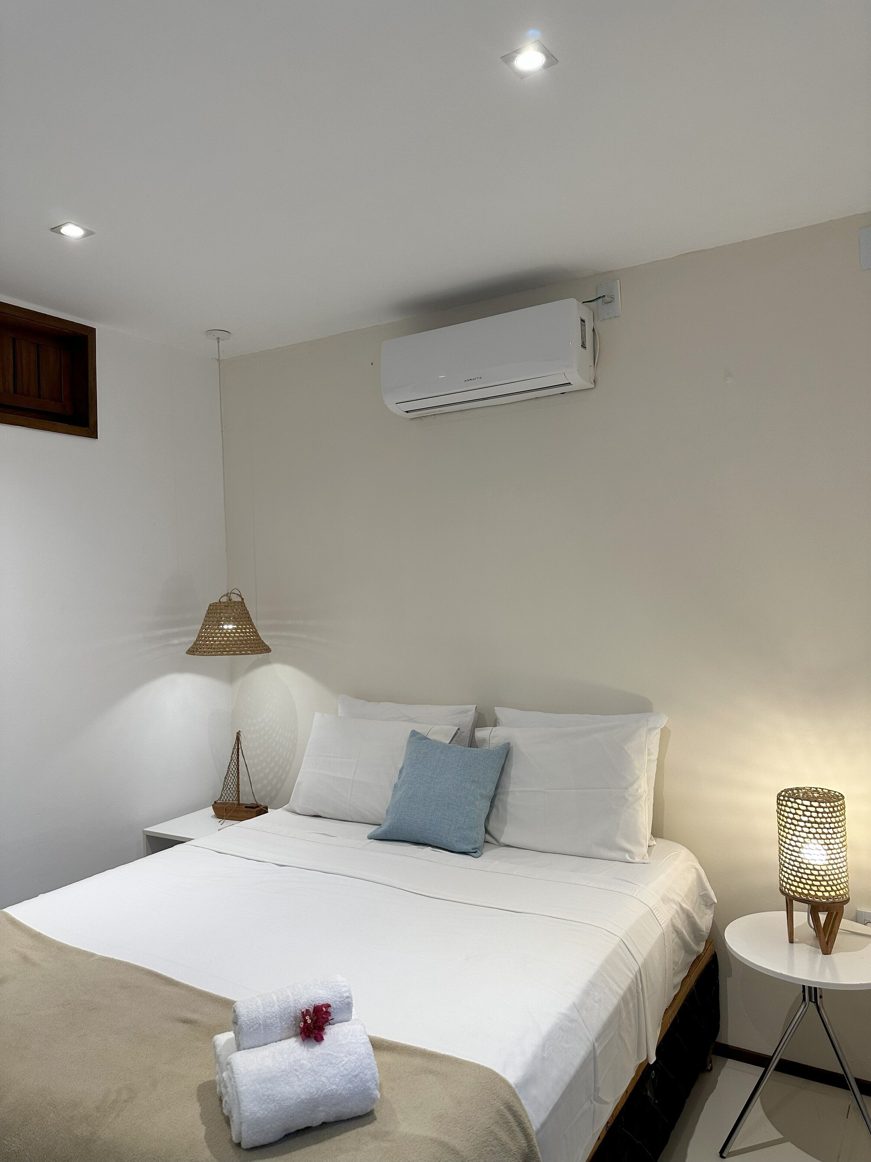 En pænt redt seng med hvide hør og en blå pude, et lille sengebord med en lampe, og en aircondition-enhed monteret på væggen.
