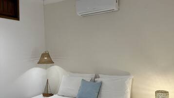 En pænt redt seng med hvide hør og en blå pude, et lille sengebord med en lampe, og en aircondition-enhed monteret på væggen.