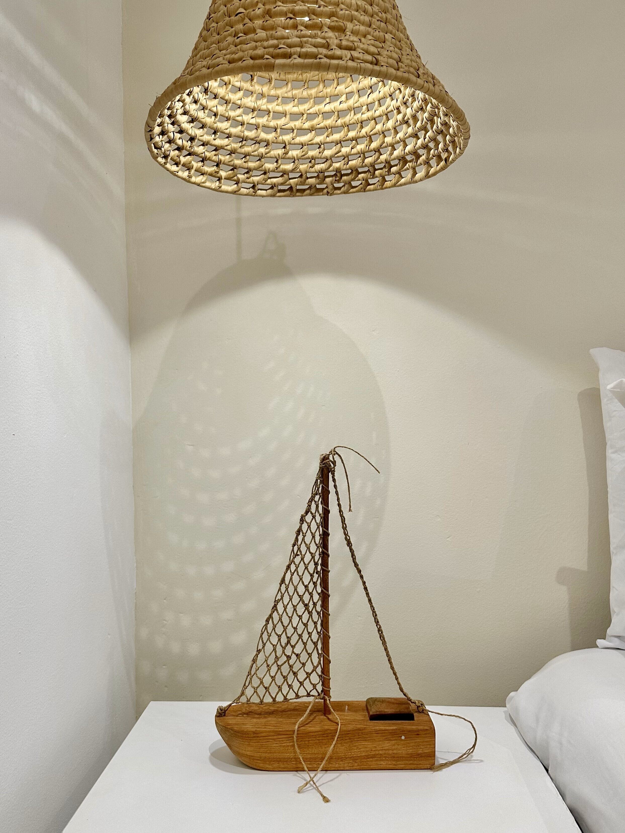 Una habitación con una cama blanca, una mesita de noche de madera, una lámpara colgante tejida y una decoración con forma de barco de madera sencillo.