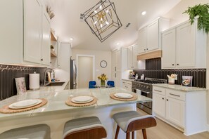 A modern kitchen with white cabinets, a central island with bar stools, a stainless steel stove, and a hanging light fixture.