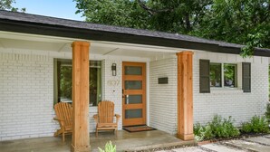A white brick house with a wooden porch, two wooden chairs, and a door with a window.