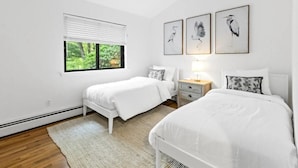 Un dormitorio con dos camas, una ventana con persianas, una mesita de noche con lámpara y tres cuadros enmarcados en la pared.