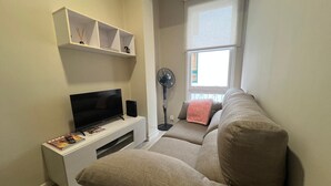 Una sala de estar con un televisor de pantalla plana sobre un pedestal blanco, un sofá beige, un estante pequeño blanco con adornos y un ventilador de pie.
