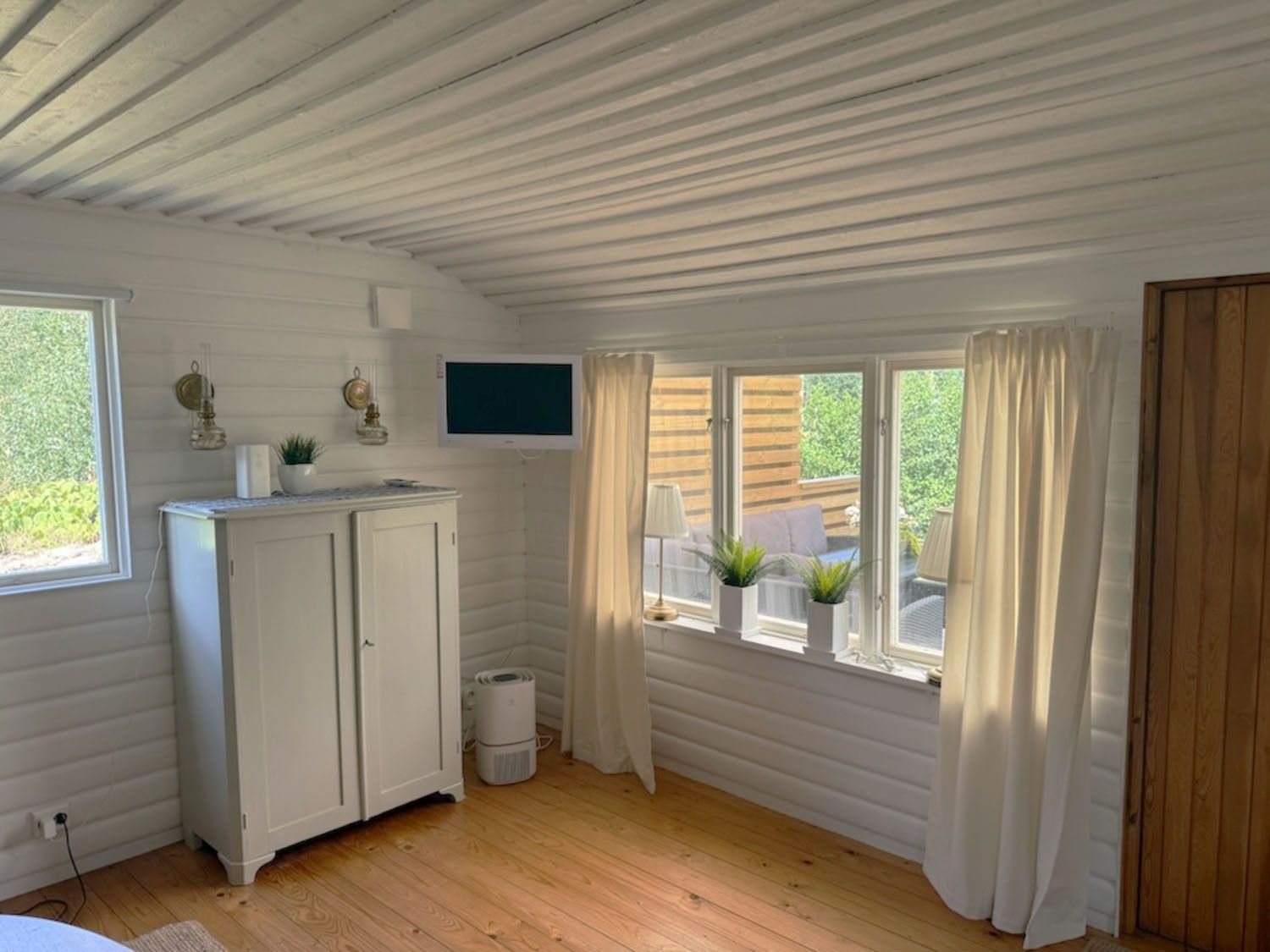 Una habitación con techo de madera, paredes blancas, un armario blanco, un televisor de pantalla plana y una ventana con cortinas.