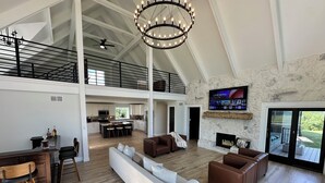 A spacious living area with a high ceiling, a fireplace, a TV mounted on the wall, and a view of the outdoors.