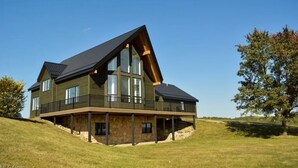 A modern two-story house with a large balcony, situated on a grassy hill.