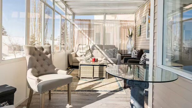 A sunlit room with large windows, a tufted armchair, a sofa, and a glass coffee table. - Sunset View House (families) (Calgary)