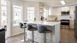 A modern kitchen with white cabinets, stainless steel appliances, and a central island with bar stools. - Sunset View House (families) (Calgary)