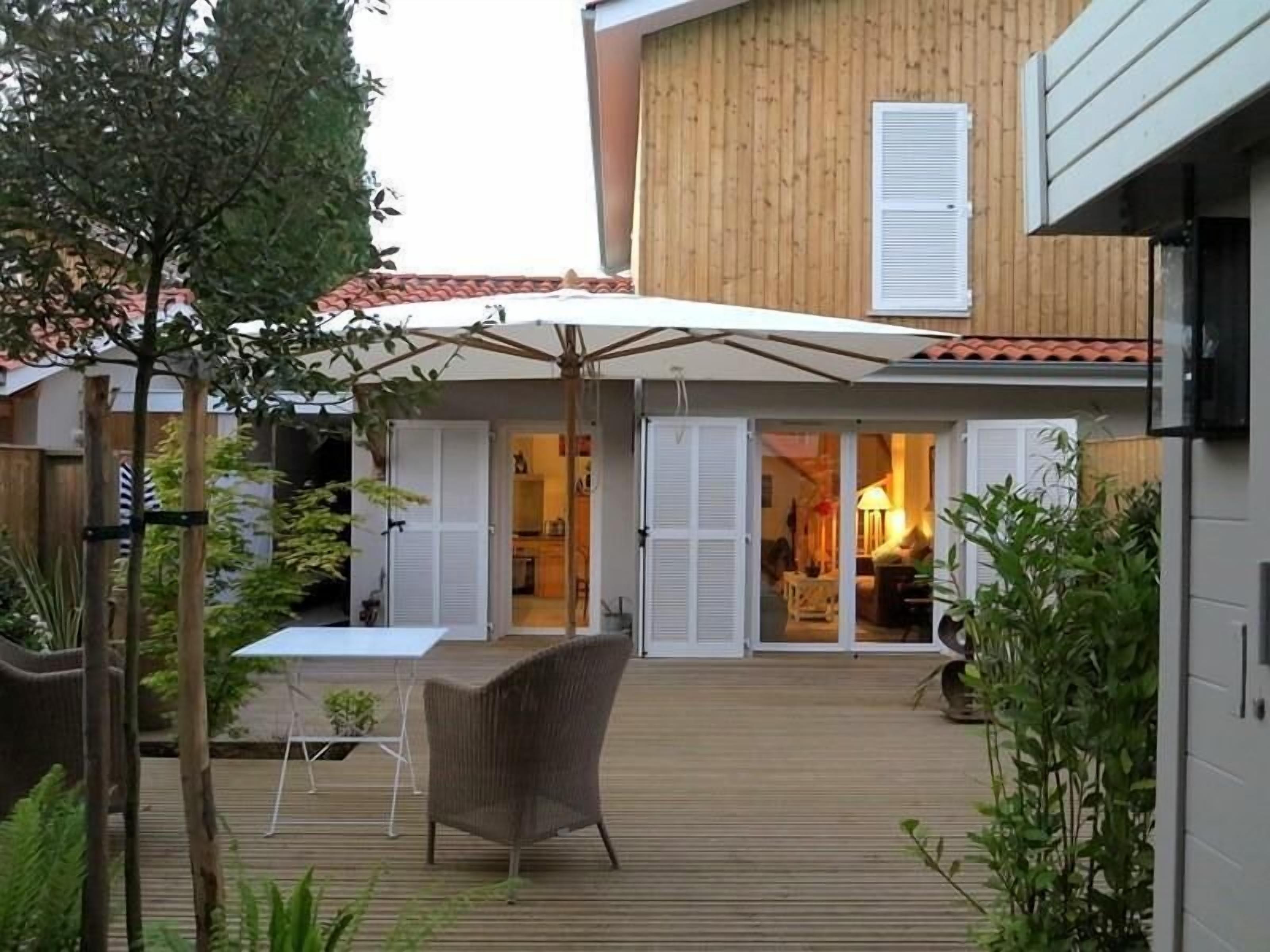 Una terraza de madera con mobiliario exterior y un porche cubierto, con vista al interior de una vivienda.