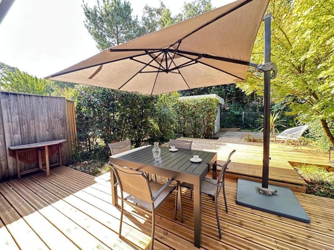 En udendørs patio med et bord og stole, en træterrasse og en stor parasol, der giver skygge.