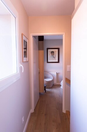 A narrow hallway with wooden flooring leading to a room with a wicker chair and a framed picture on the wall.