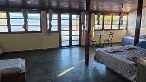 Una habitación con cama, una mesa y sillas, ventanas amplias con cortinas y vistas al mar.