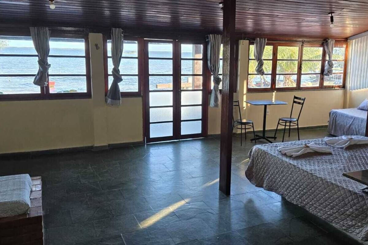 Una habitación con cama, una mesa y sillas, ventanas amplias con cortinas y vistas al mar.