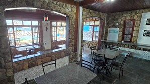 Un comedor de paredes de piedra con mesas y sillas, ventanas amplias y vistas al exterior.