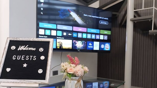 Un espace d’accueil doté d’un écran numérique affichant diverses icônes d’applications, d’une pancarte de bienvenue pour les visiteurs, d’un vase avec des fleurs et d’une télécommande posée sur une table en verre.