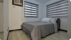 Une chambre à coucher avec un lit, deux fenêtres équipées de stores, un climatiseur fixé au mur et une prise de courant.
