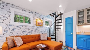 A room with a sofa, a small round table, a staircase, and a kitchen area with blue cabinets. - Tiny House Living + Parking | Near West End Beach (Provincetown)