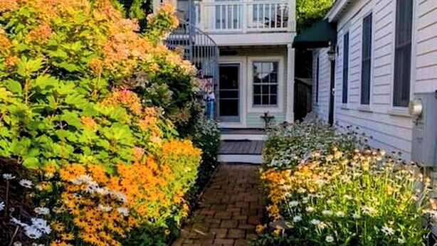 A two-story house with a front porch, surrounded by a garden with various plants and flowers. - Tiny House Living + Parking | Near West End Beach (Provincetown)
