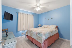 Quarto com cama, ventilador de teto, televisão, janela com cortinas e mesa de cabeceira com abajur.
