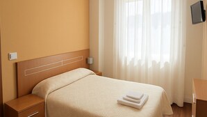 Ένα δωμάτιο ξενοδοχείου με ένα ξύλινο κρεβάτι, ένα κομοδίνο, ένα φωτιστικό δίπλα στο κρεβάτι, μια τηλεόραση επίπεδης οθόνης και ένα μεγάλο παράθυρο με λεπτά, διάφανα κουρτίνια.
