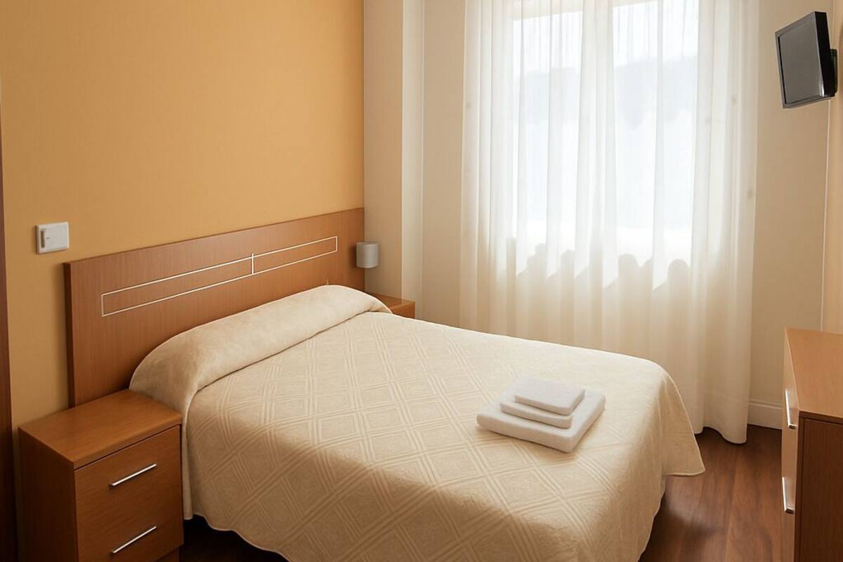 A hotel room with a wooden bed, a nightstand, a bedside table, a flat-screen TV, and a large window with sheer curtains.