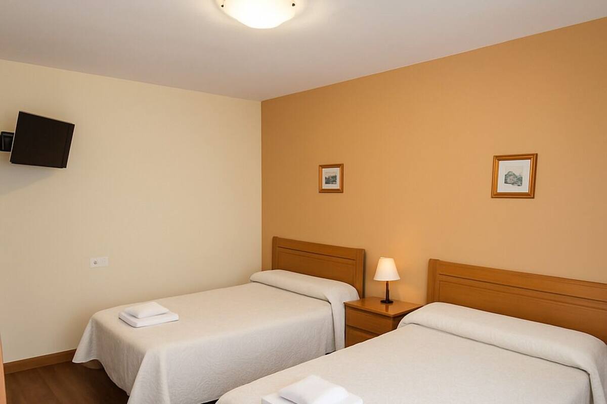 A hotel room with two single beds, a bedside table with a lamp, a flat-screen TV mounted on the wall, and two framed pictures on the wall.