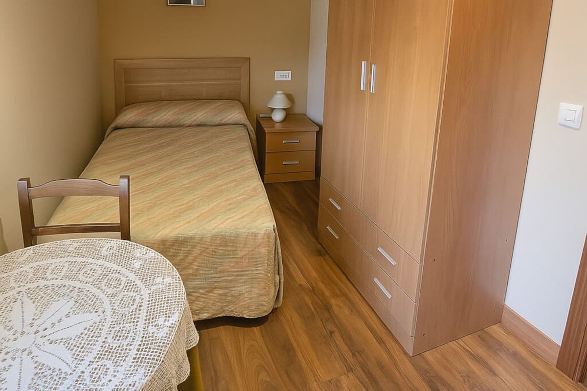 A bedroom with a bed, a wooden wardrobe, a bedside table with a lamp, and a small table with a lace tablecloth.