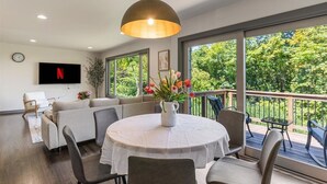 A modern living room with a round table, comfortable seating, a large window with a view of greenery, and a mounted TV.