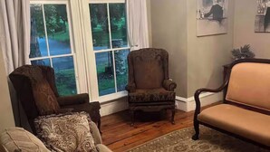 A living room with a brown sofa, a patterned armchair, a beige sofa, a wooden floor, a large window with curtains, and a chandelier.