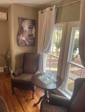 Una habitación con piso de madera, un sillón marrón oscuro, una mesa redonda con un tablero de ajedrez, una obra de arte de una guitarra en la pared, y una ventana con cortinas.