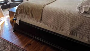 A bedroom with a large bed, a wooden floor, a rug, a ceiling light, and a window with curtains.