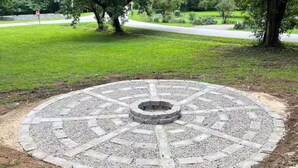 A circular stone patio with a central fire pit, surrounded by a grassy park area with trees and a road in the background.