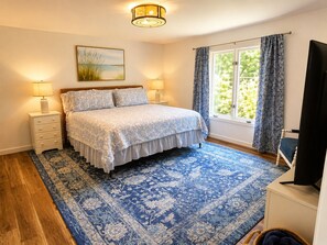 Quarto com cama, duas luminárias de cabeceira, janela com cortinas, um quadro na parede e uma televisão sobre um suporte.