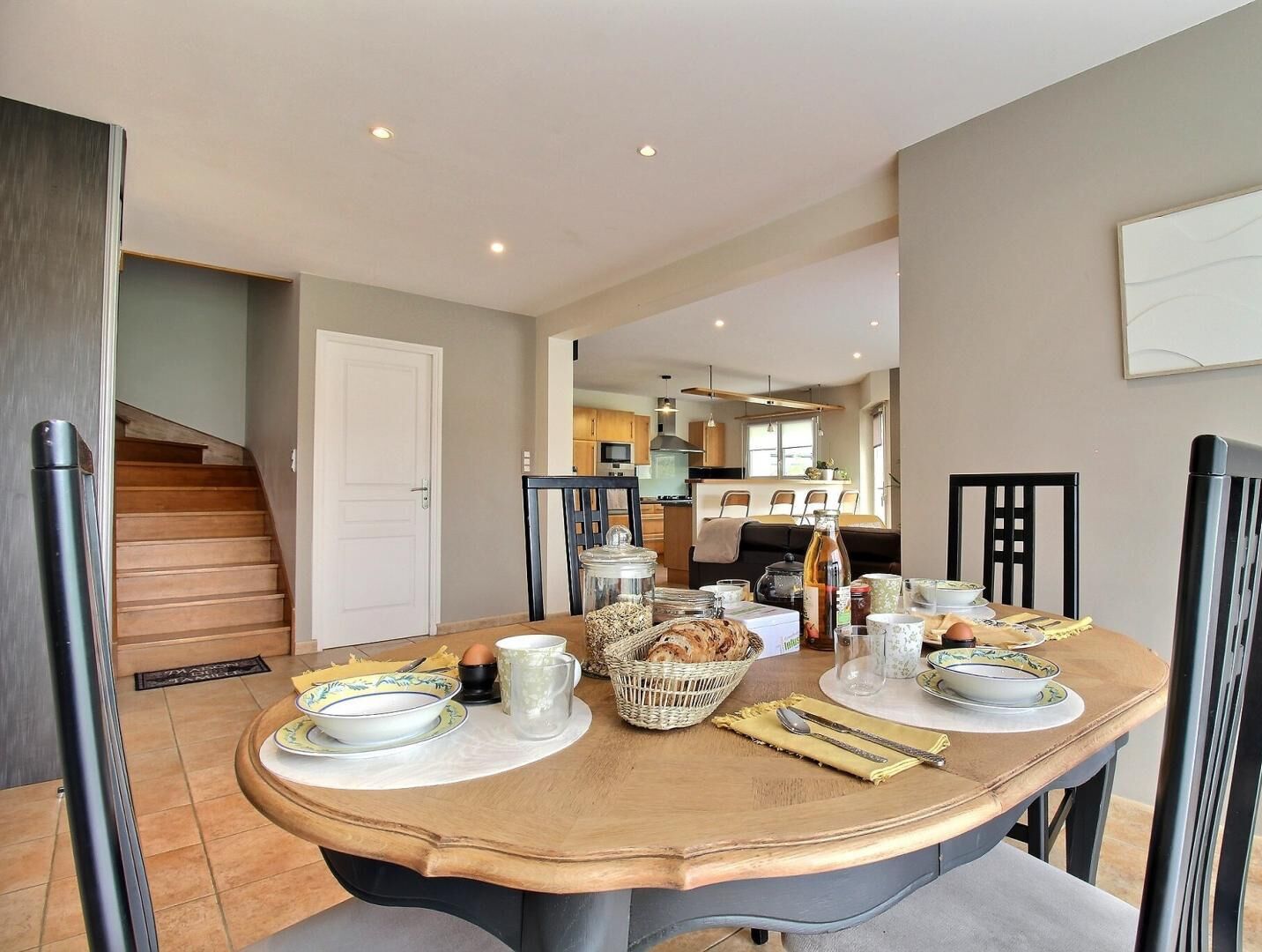 A dining area with a wooden table set for four, surrounded by chairs. There is a kitchen in the background with modern appliances and a dining table.
