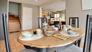 A dining area with a wooden table set for four, surrounded by chairs. There is a kitchen in the background with modern appliances and a dining table.