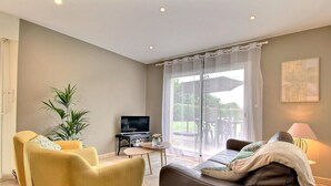 A living room with a sofa, armchairs, a TV, and a table. There is a large window with curtains and a view of the outdoors.