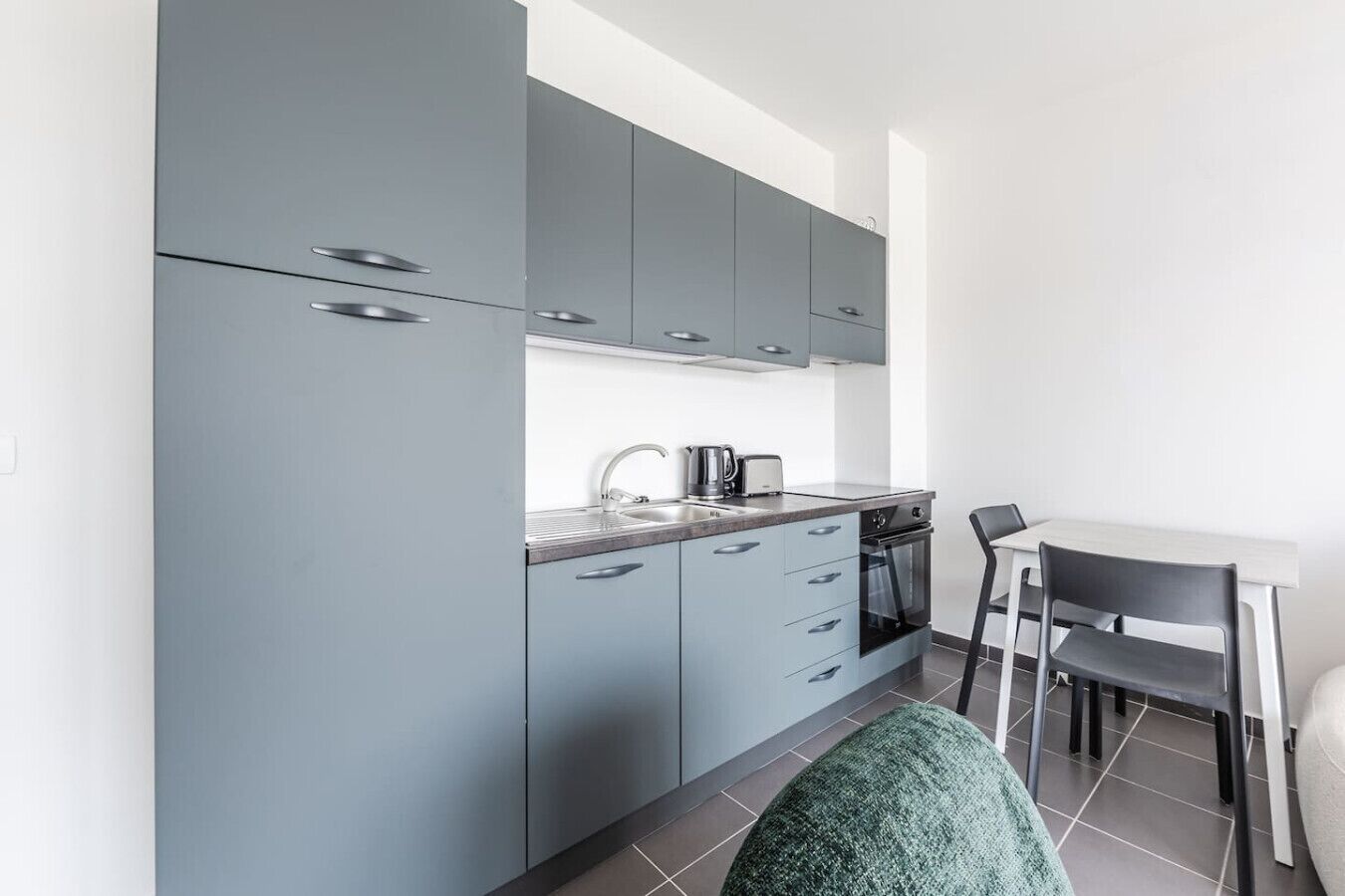 Een moderne keuken met grijze kastjes, een spoelbak, een waterkoker en een fornuis. Er staat een eettafel met twee stoelen.
