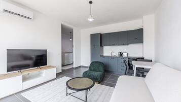 A modern living room with a flat-screen TV, a sofa, and a small round table. There is a kitchen area with grey cabinets and a dining table.