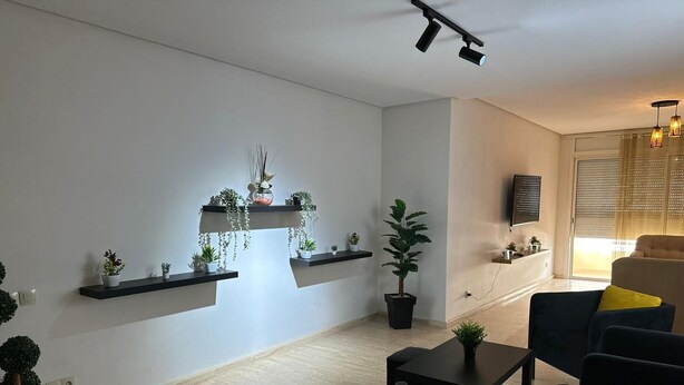 A modern living room with a sofa, a coffee table, and a TV mounted on the wall. There are plants on floating shelves and a potted plant on the floor.