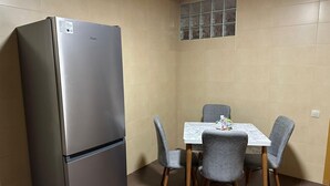 A compact kitchen with a stainless steel refrigerator, a small dining table with four grey chairs, and a tiled backsplash.